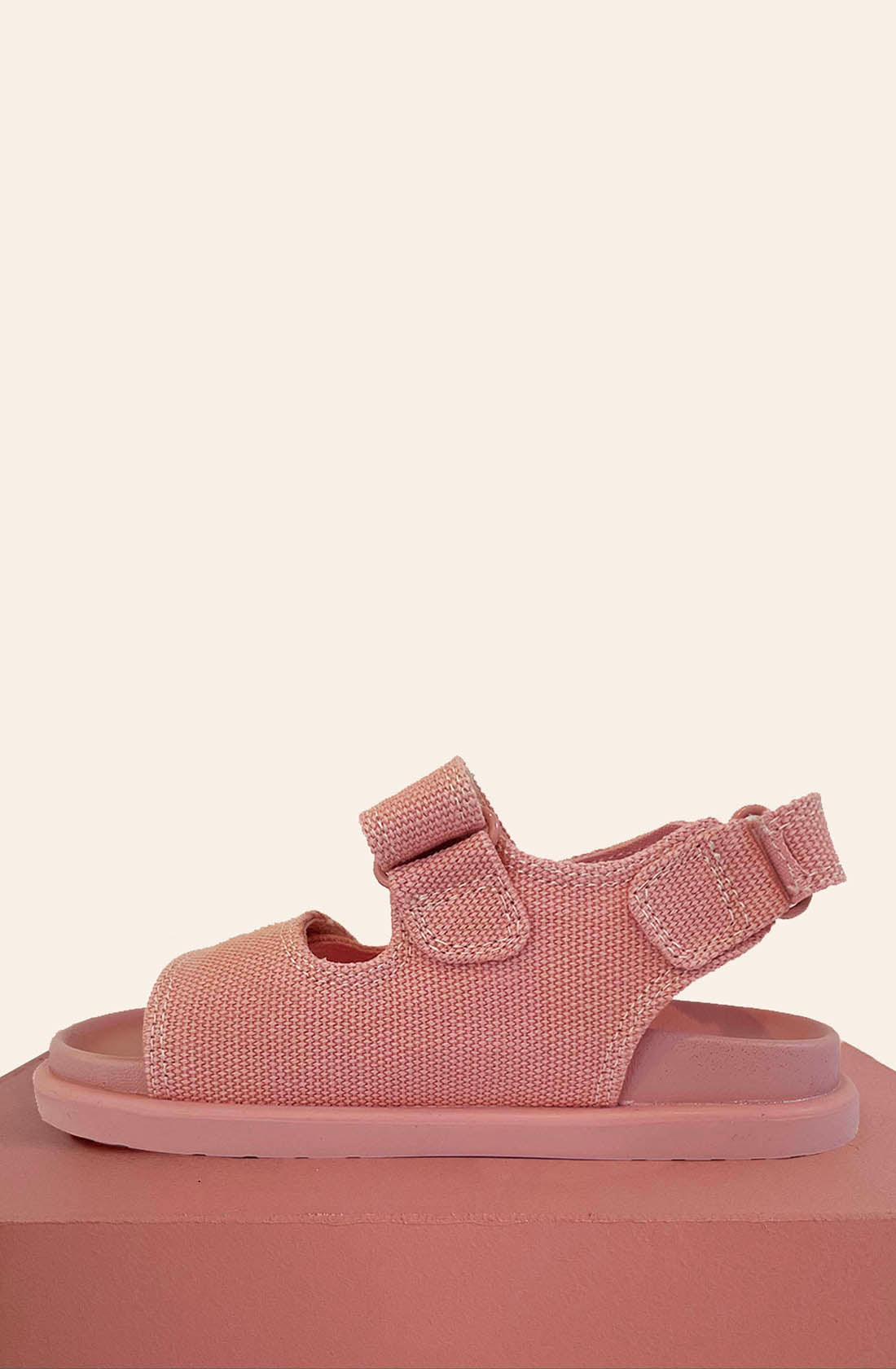 The PRE-ORDER Original Sandals Pink by PICCOLINI are showcased on a matching pink block against a cream backdrop. These children's sandals feature adjustable Velcro straps, a textured fabric upper, and a non-slip rubber sole for both safety and style.