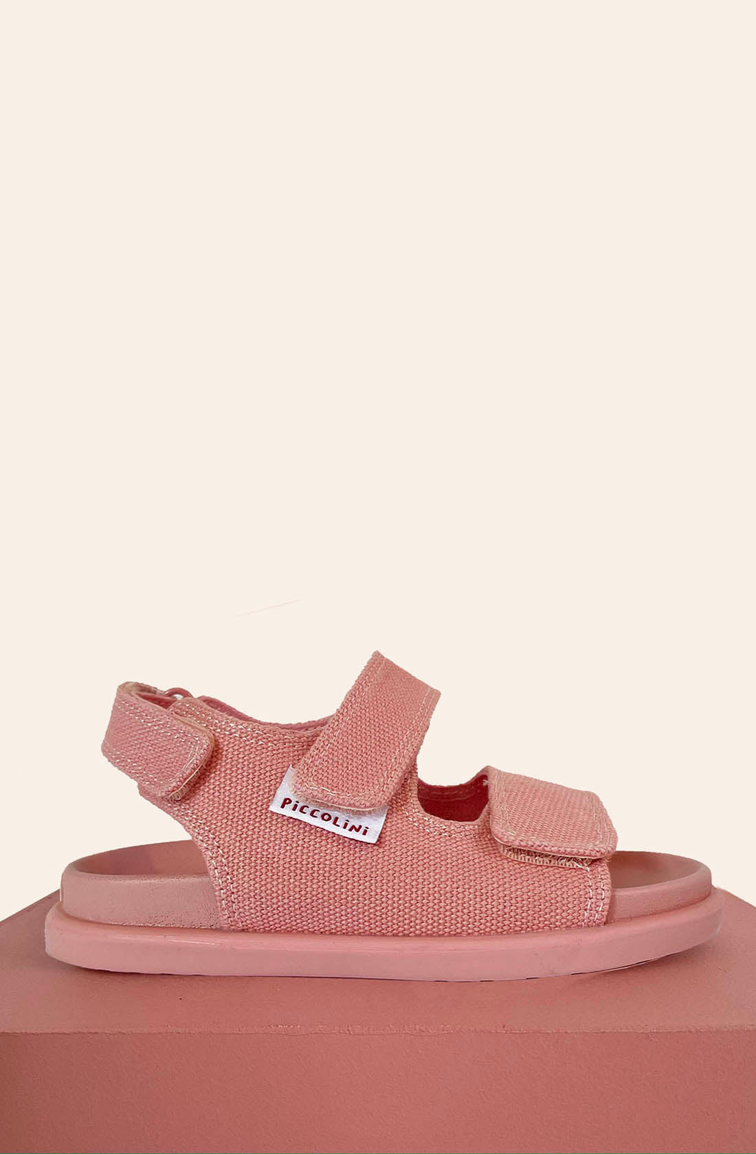 The PRE-ORDER Original Sandals Pink by PICCOLINI, designed as pink children's sandals with Velcro straps, a non-slip rubber sole, and a small label on the side, are displayed on a pink cube set against a beige background.