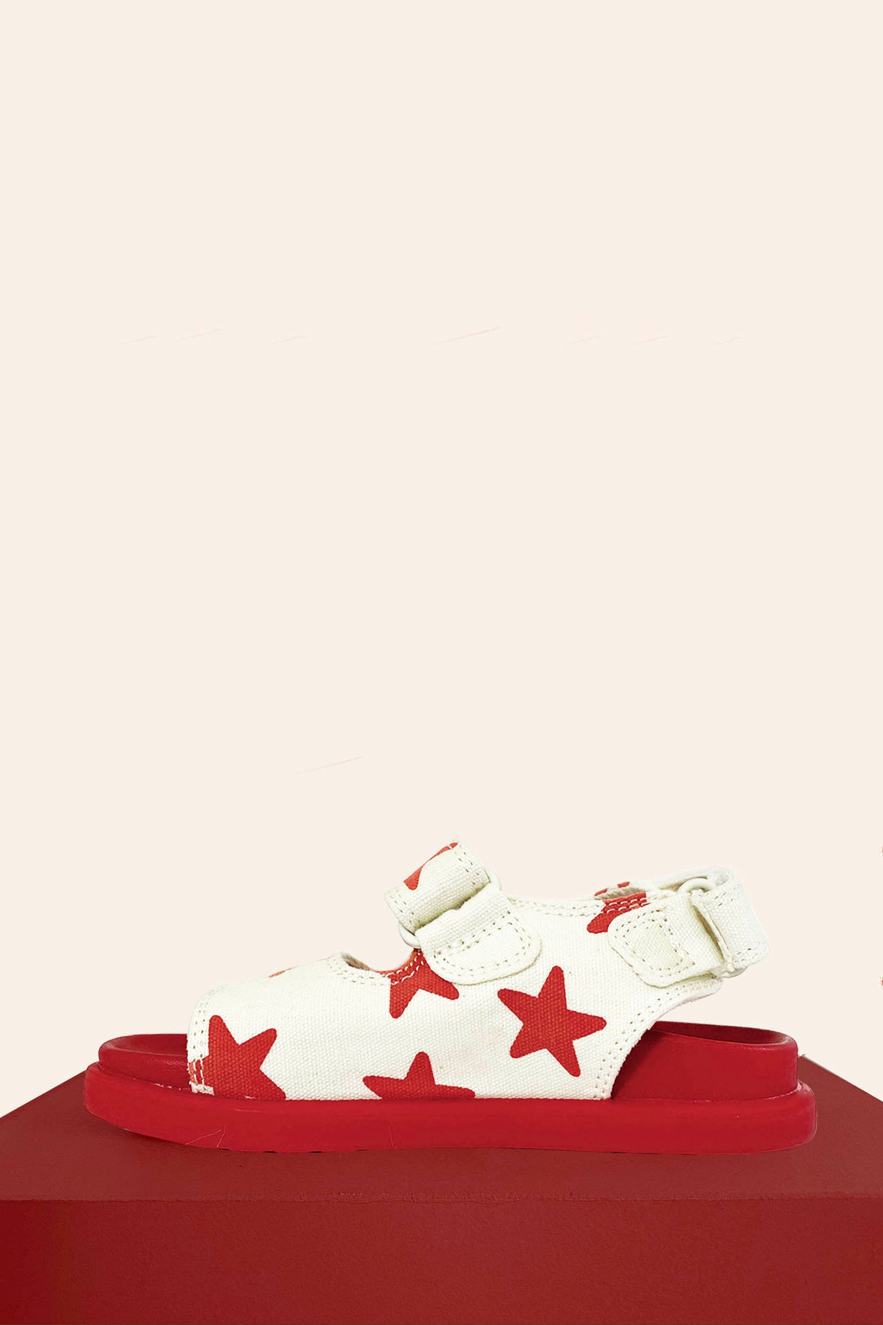 The PRE-ORDER Limited Edition Piccolini x By Billie Sandals Red Star by PICCOLINI, featuring a children's sandal design with a white upper adorned with red stars, an adjustable strap, and a non-slip rubber sole, is displayed on a red surface against a plain cream background.