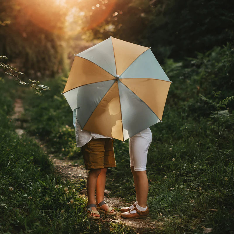 Two people standing on a narrow path in a lush, green forest share a large See-Ya Umbrella from OLLI ELLA made from recycled polyester that obscures their faces. The scene is lit by a warm, golden light from above, creating a romantic and idyllic atmosphere.