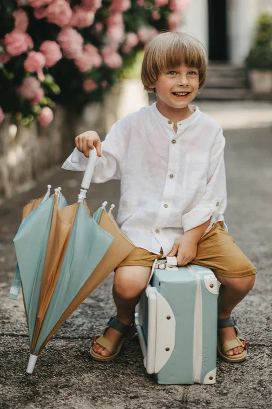 A young boy with blond hair is sitting on a mint green suitcase, wearing a white shirt, tan shorts, and sandals. He is holding a See-Ya Umbrella from OLLI ELLA made from recycled polyester with blue and tan panels. He smiles while sitting on a pathway surrounded by pink flowers.