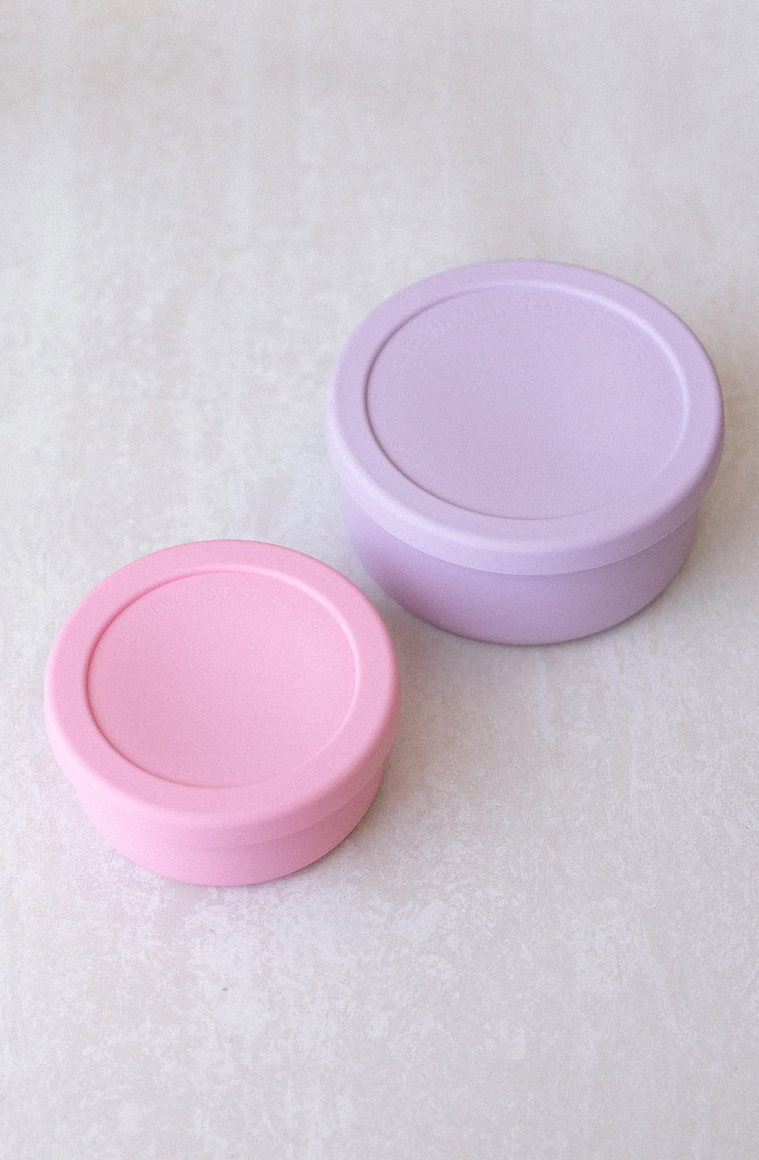 Two round, BPA-free silicone containers from THE WHOLESOME STORE are placed on a light-colored surface. The larger, 700ml container is lavender, and the smaller one is pink. Both containers feature a simple, minimalist design and are dishwasher safe for easy cleaning.