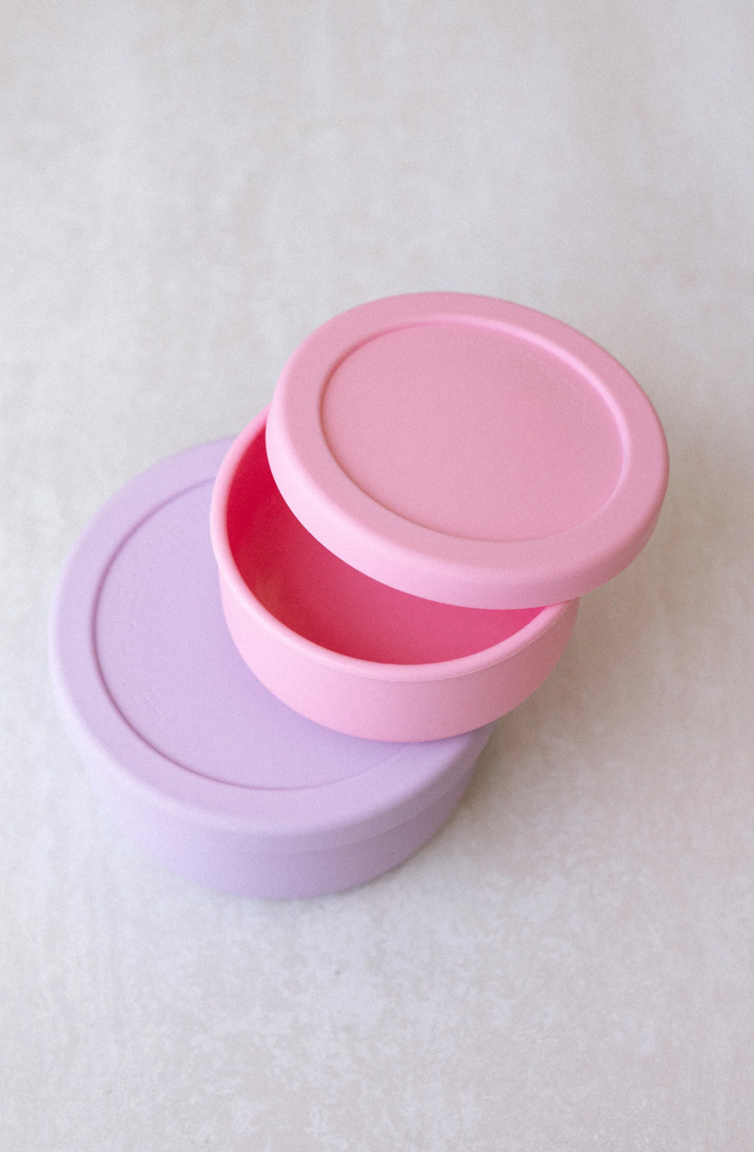 Two circular silicone Round Silicone Containers Small (400ml) from THE WHOLESOME STORE, one pink and one lavender, are stacked on a light surface. The pink container's lid is slightly open, revealing its empty interior. The lavender container is fully closed beneath it. Both BPA-free containers are also dishwasher safe for easy cleaning.