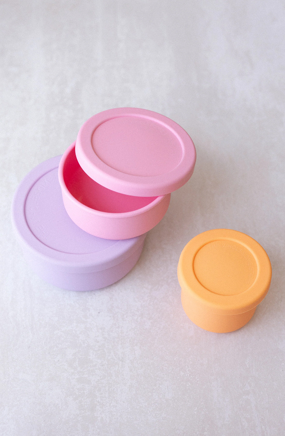 Three round, colorful BPA-free containers from THE WHOLESOME STORE's Silicone Snack Pot 250ml collection are stacked on a light surface. The largest is pastel purple, the middle is pink, and the smallest, separate from the stack, is orange. The lids are on each container except for the pink one, which is slightly askew. These silicone round snack pots are also dishwasher safe.