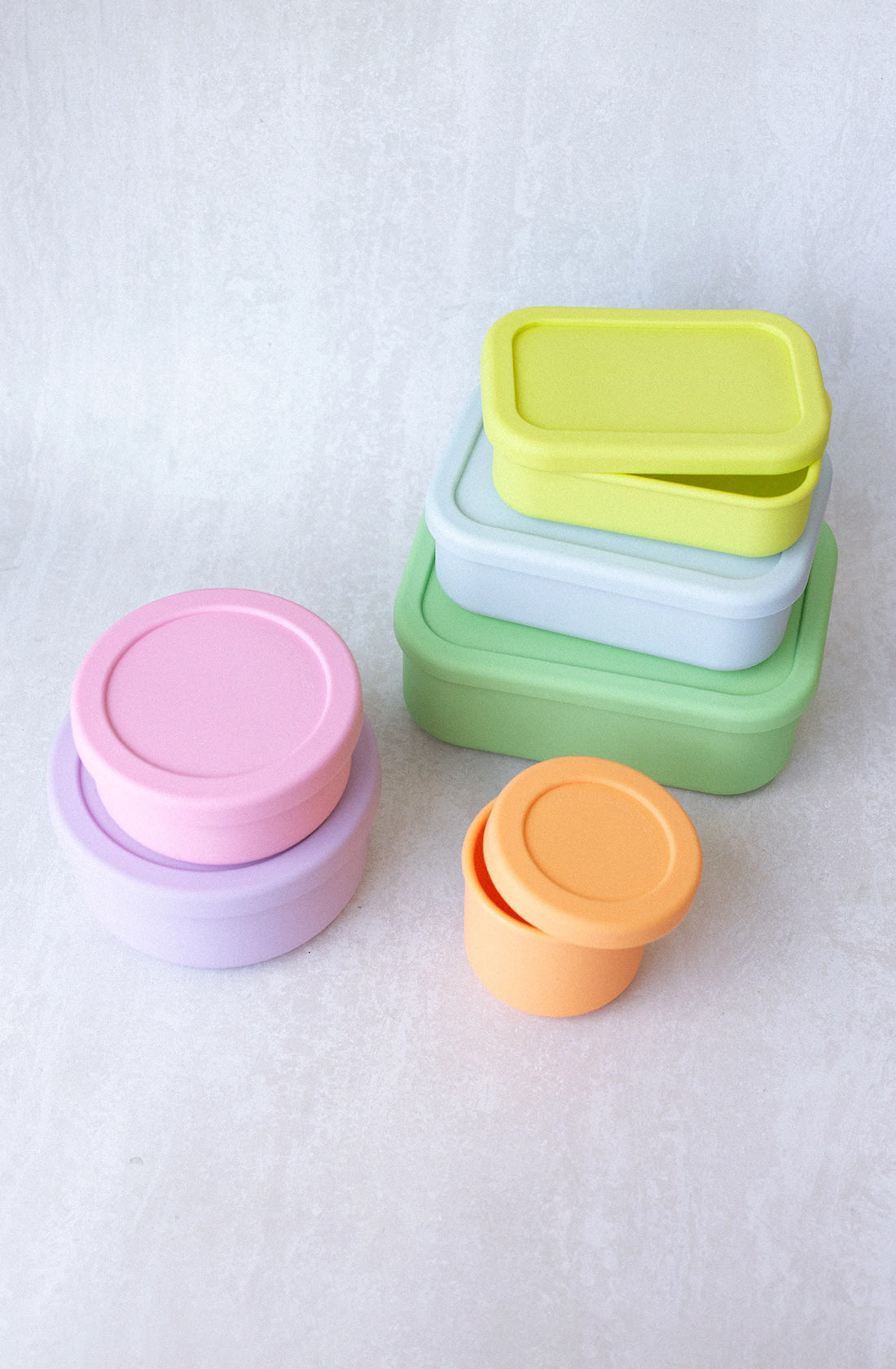 A set of colorful, stackable BPA-free Silicone Snack Boxes 300ml from THE WHOLESOME STORE are arranged neatly on a light gray surface. These snack boxes come in various pastel colors, including pink, purple, orange, green, and yellow, and in different shapes and sizes. Plus, they are dishwasher safe for easy cleaning.