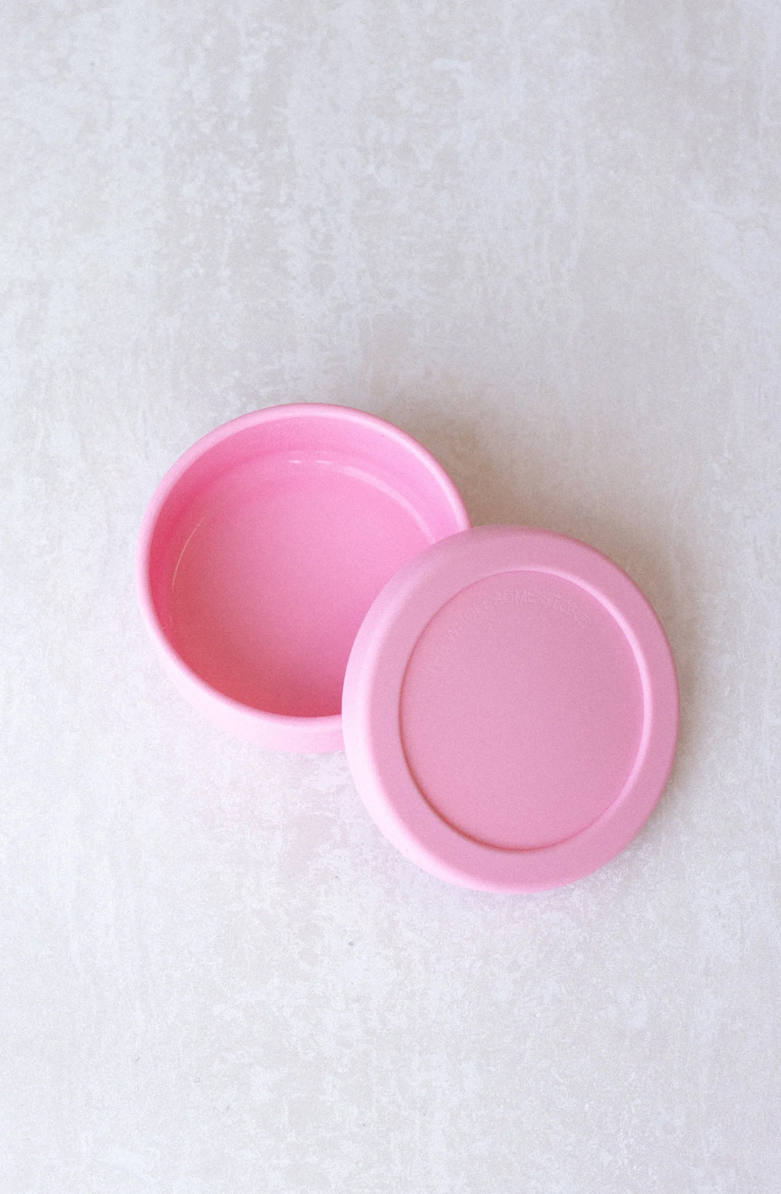 A pink, circular container from THE WHOLESOME STORE labeled as the Round Silicone Container Small (400ml) with a lid placed beside it on a light textured surface. This BPA-free container appears to be empty and is conveniently dishwasher safe.