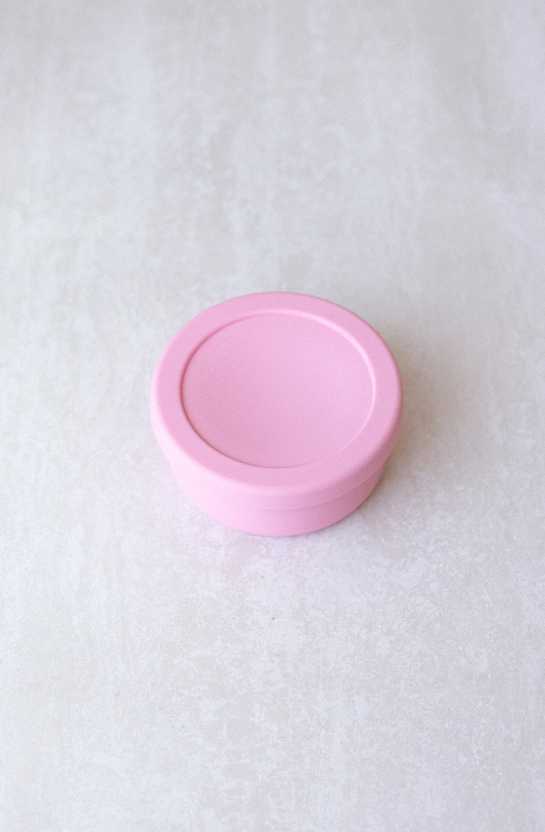 A small, round, pastel pink container from THE WHOLESOME STORE, complete with a matching lid, is set against a light grey, slightly textured background. This 400ml Round Silicone Container Small appears to be both BPA-free and dishwasher safe, combining practicality with style.