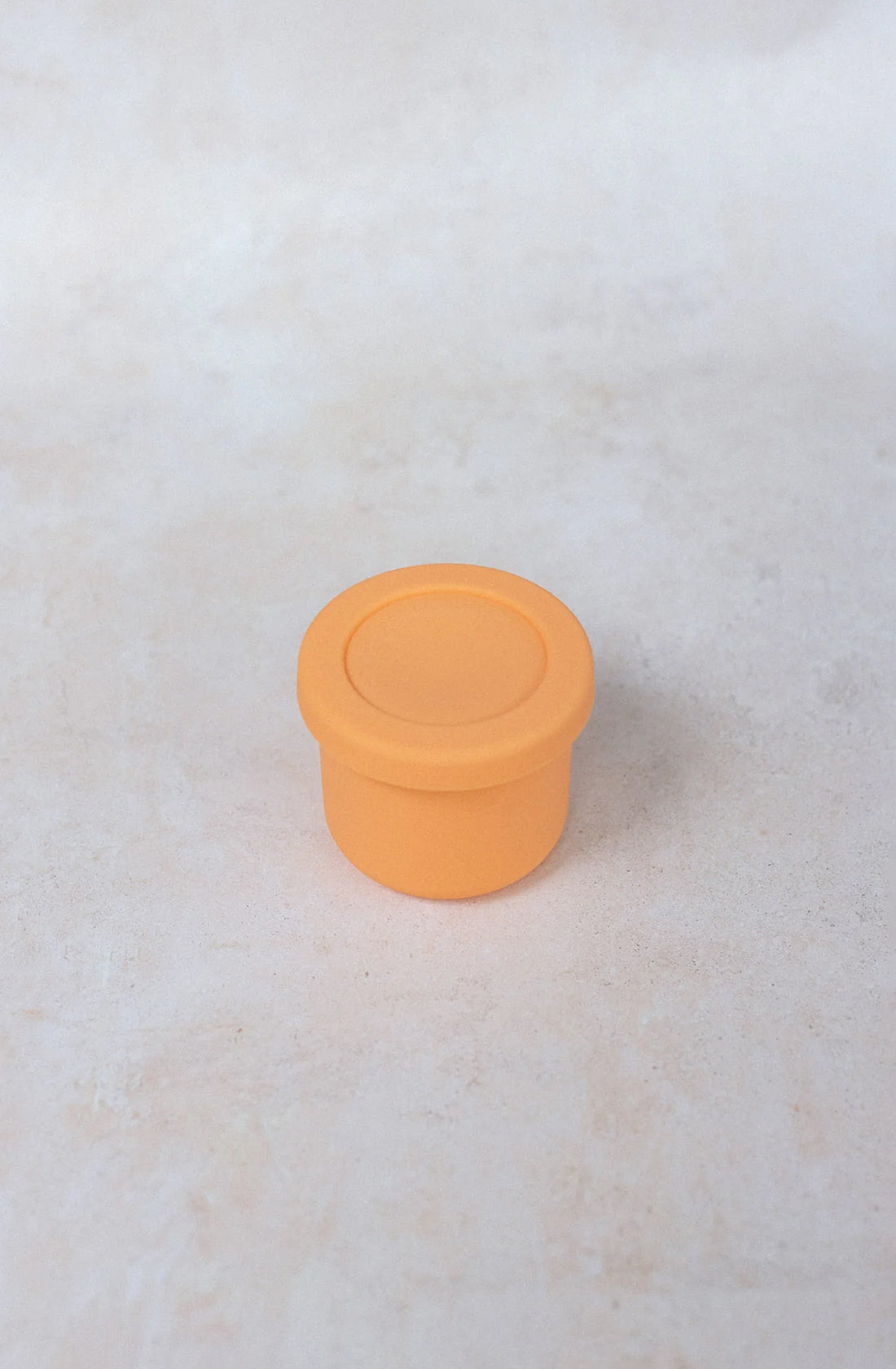 A small, rounded BPA-free container with a flat top, made of smooth, light orange silicone material, is placed on a neutral, light-colored surface. The Silicone Snack Pot 250ml by THE WHOLESOME STORE appears to be simple and minimalistic in design and is also dishwasher safe.