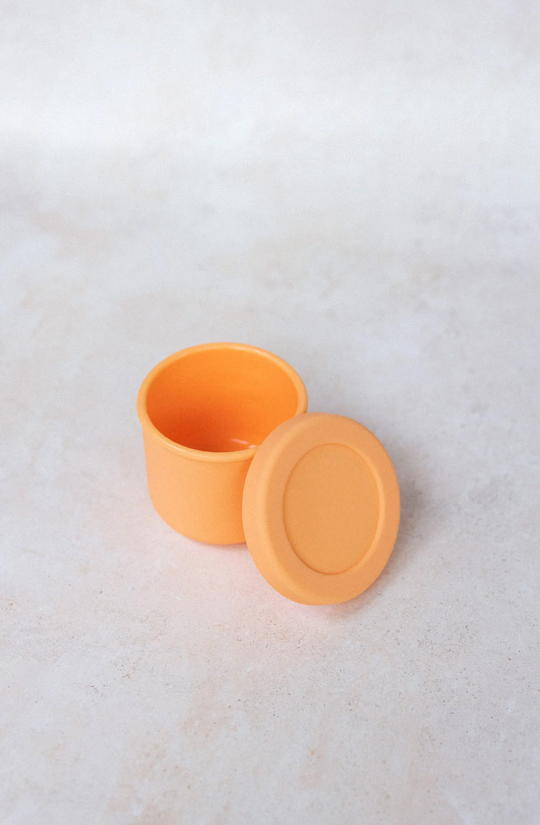 A small, round, orange BPA-free container from THE WHOLESOME STORE sits with its matching lid beside it. The Silicone Snack Pot 250ml is open, revealing its empty interior against a light, textured background. This convenient snack pot is also dishwasher safe.