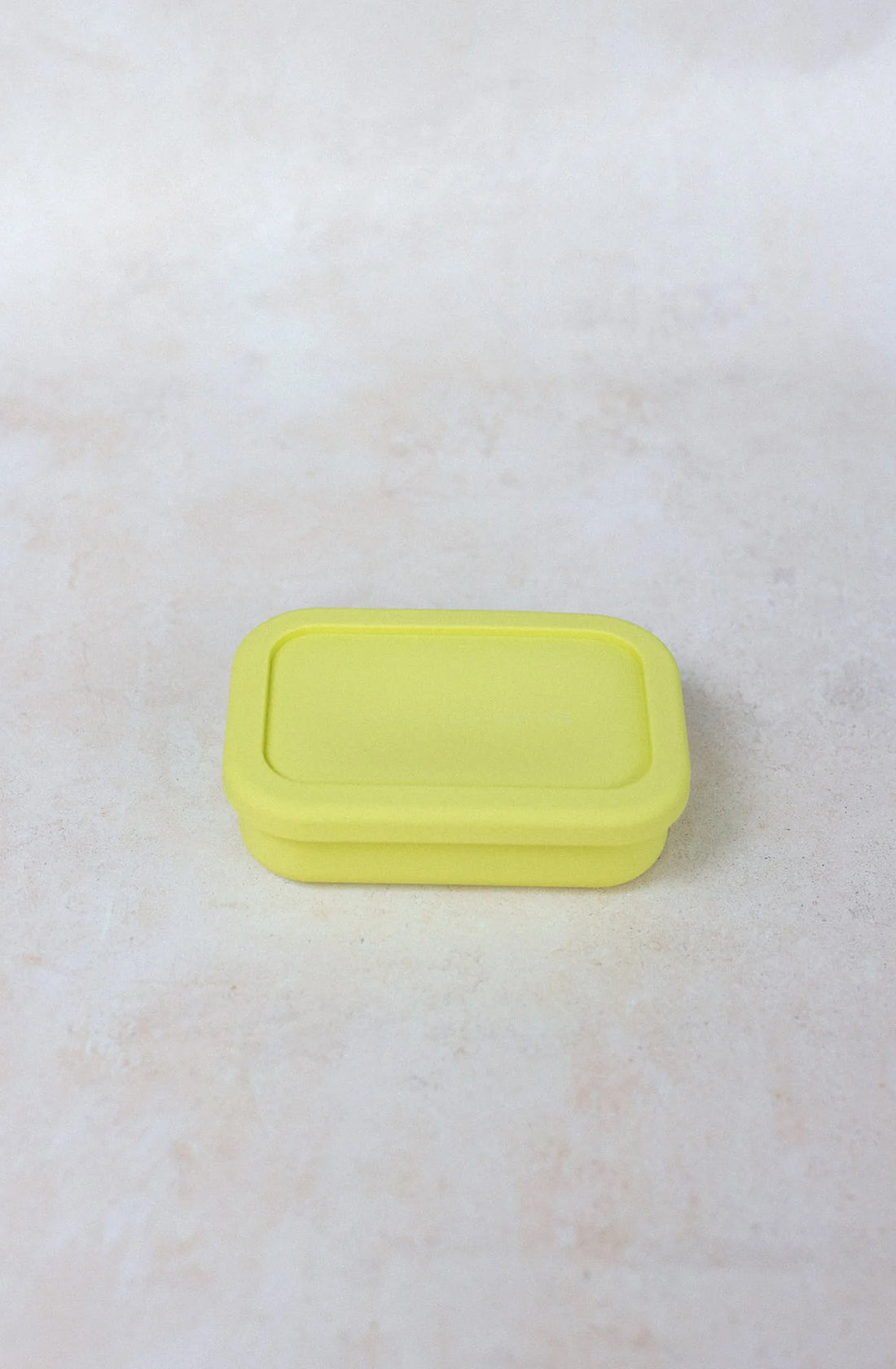 A small, rectangular, yellow BPA-free container with a lid, the Silicone Snack Box 300ml from THE WHOLESOME STORE, sits on a light cream-colored surface. Its simple, unadorned design makes it perfect for any kitchen.