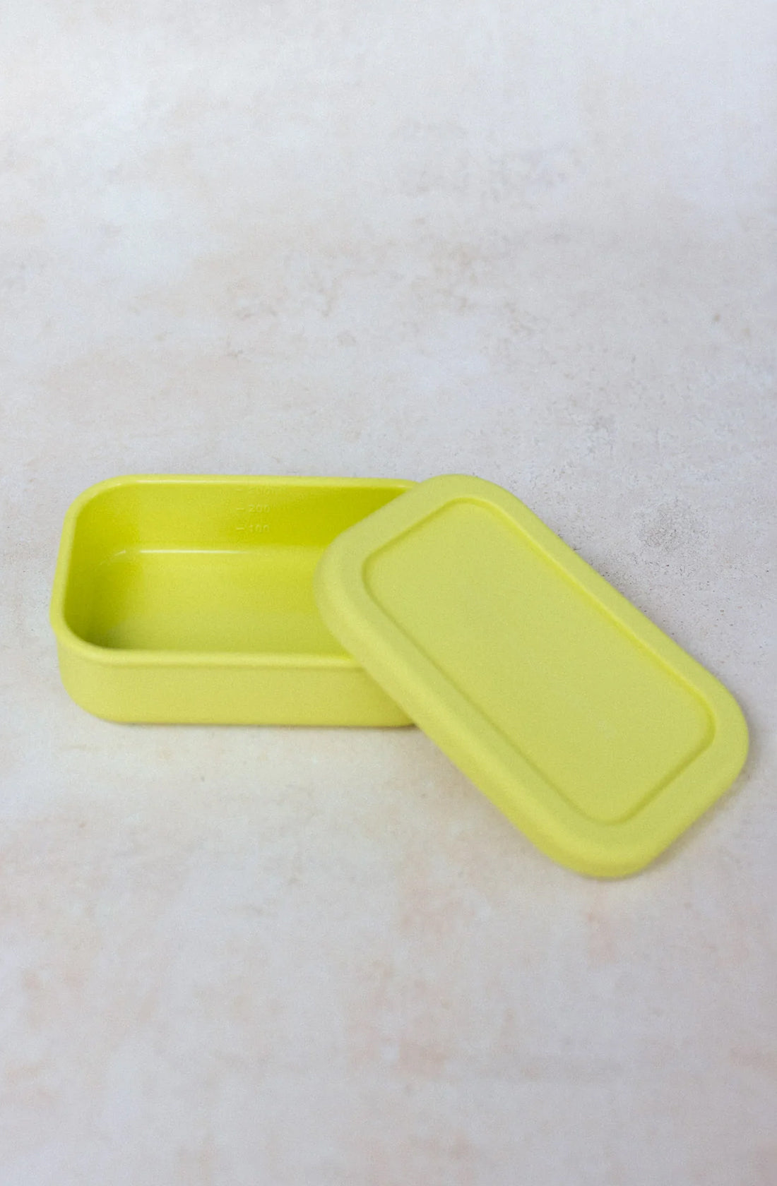 A light green rectangular Silicone Snack Box 300ml from THE WHOLESOME STORE sits on a beige surface. Its matching lid is removed and placed slightly angled to the right, next to the container. The simple design is minimalist with smooth edges and is BPA free.