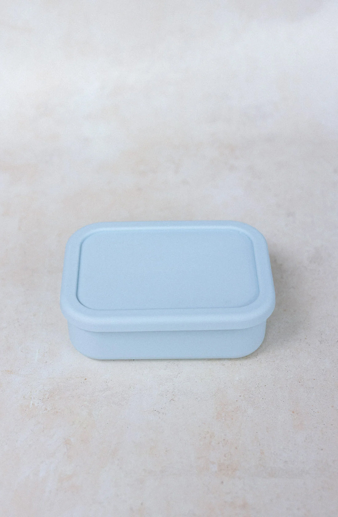 A light blue silicone Bento Lunchbox Small (700ml) from THE WHOLESOME STORE, featuring a fitted lid, smooth edges, and a minimalist design, is placed on a beige surface. This BPA-free container combines practicality with style.