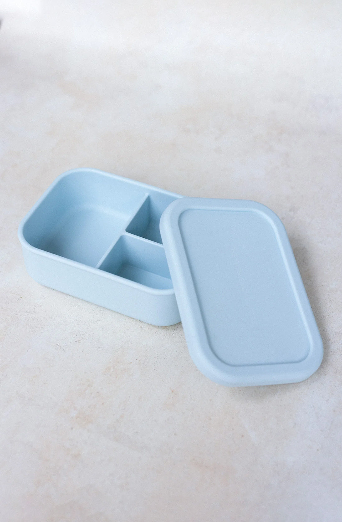 A Silicone Bento Lunchbox Small (700ml) by THE WHOLESOME STORE in light blue with a matching lid is shown open. This BPA-free lunchbox features three compartments: one large and two smaller ones, and it is placed on a light-colored, textured surface.