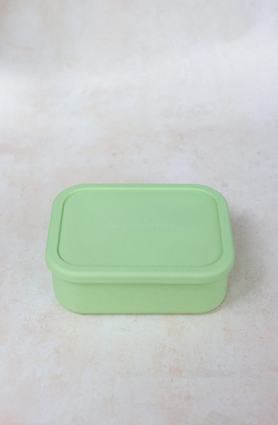 A rectangular light green plastic container from THE WHOLESOME STORE, the Silicone Bento Lunchbox Large (1300ml), with a matching lid is placed on a flat, light-colored surface. The lunchbox appears to be empty and is likely used for storage or packing food. The dishwasher-safe design ensures convenience, while its airtight cavities keep contents fresh.