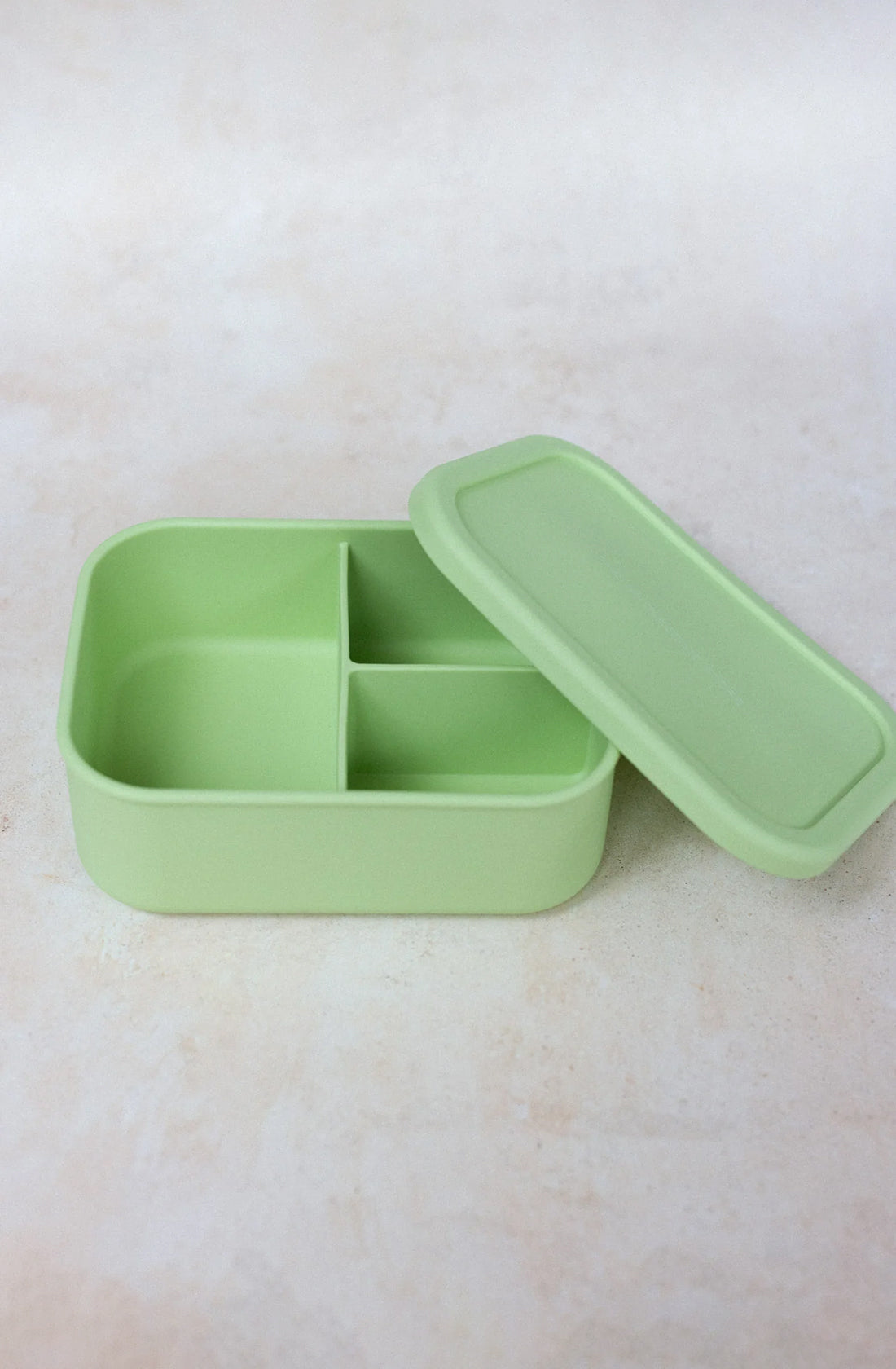 A light green "Silicone Bento Lunchbox Large (1300ml)" by THE WHOLESOME STORE, featuring three airtight compartments and a matching lid, sits next to it on a light, textured surface. The lunchbox is also dishwasher safe.