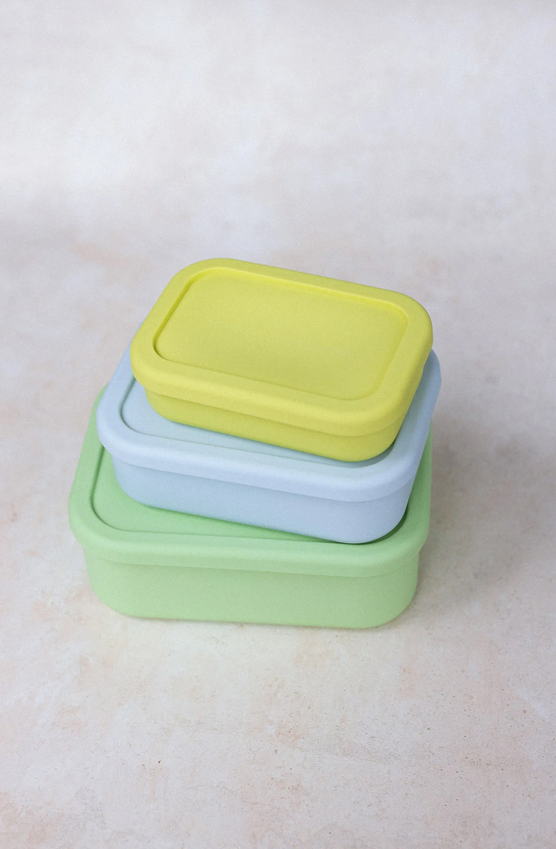 Three rectangular food storage containers, featuring airtight cavities, are stacked on top of each other. The top container is yellow, the middle one is light blue, and the bottom one is light green. They sit on a smooth, light beige surface and are dishwasher safe for easy cleaning. These are Silicone Bento Lunchboxes (1300ml) from THE WHOLESOME STORE.