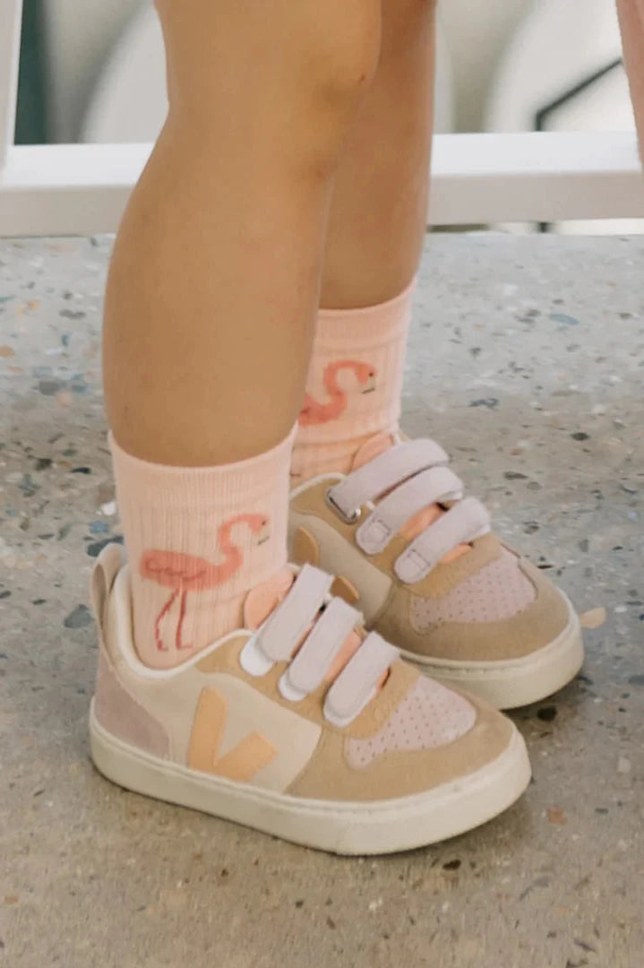 A child wearing GOLDEN CHILDREN's Flamingo Socks Sunset Pink and beige sneakers with reinforced toe caps stands on a speckled floor.