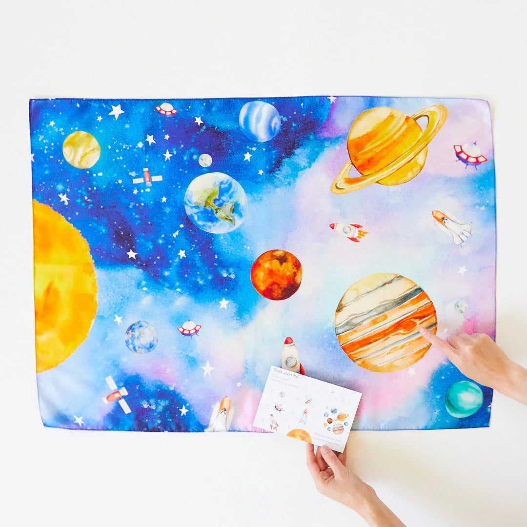 Designed for open-ended play, the SARAH'S SILKS Outer Space Seek & Find Playsilk features a colorful space theme with planets, rockets, and stars. A person's hands point to a planet while holding a card from the collection that showcases similar imaginative illustrations.