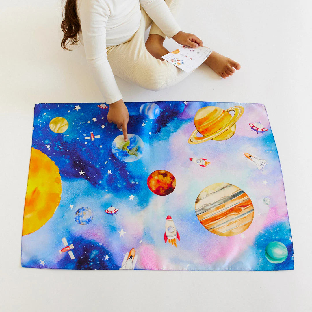 A child sits on the floor, pointing to the Outer Space Seek & Find Playsilk by SARAH'S SILKS, which features a vibrant depiction of planets, rockets, and stars on a colorful blue mat. The child's eyes light up as they hold a piece of paper featuring similar designs, engaging in open-ended play that fuels endless curiosity.