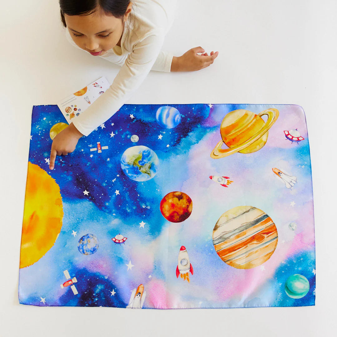 A child wearing a white shirt excitedly points at SARAH'S SILKS Outer Space Seek & Find Playsilk, which is adorned with planets, stars, rockets, and the sun. Spread out on a white surface, this imaginative playsilk inspires curiosity as the child holds a booklet filled with vibrant illustrations.