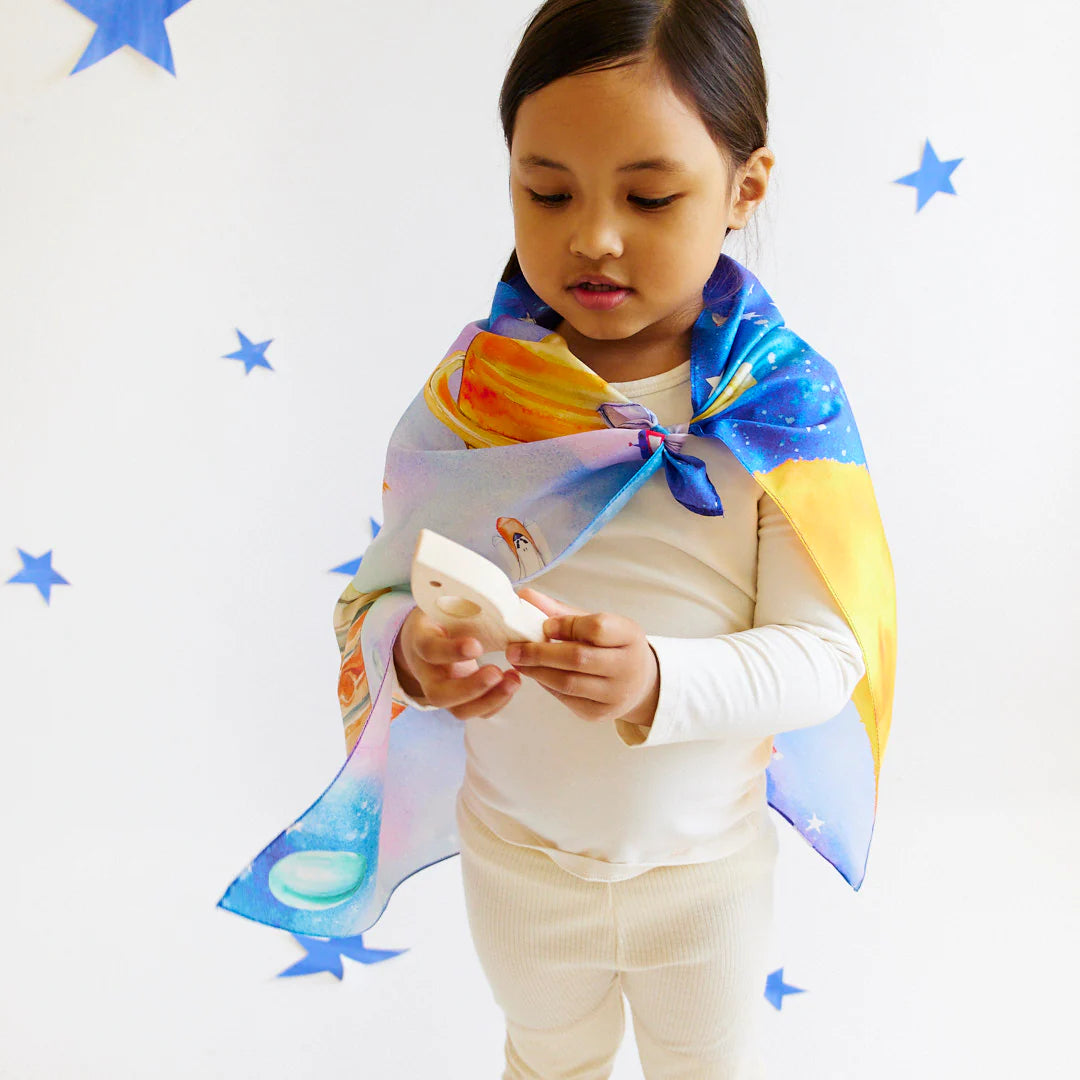 Amidst a backdrop of white adorned with blue stars, a child is enveloped in SARAH'S SILKS' Outer Space Seek & Find Playsilk, fashioned like a cape. Engaged in boundless imaginative play, they are intently observing a small toy or object cradled within the playsilk's folds.