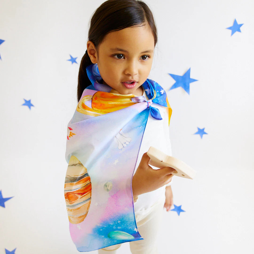 A child with long dark hair stands wearing the Outer Space Seek & Find Playsilk, a vibrant cape adorned with planets, ideal for open-ended play. The background is decorated with blue stars. Holding a wooden toy, the child gazes thoughtfully to the side, lost in their own universe. This imaginative playsilk is by SARAH'S SILKS.