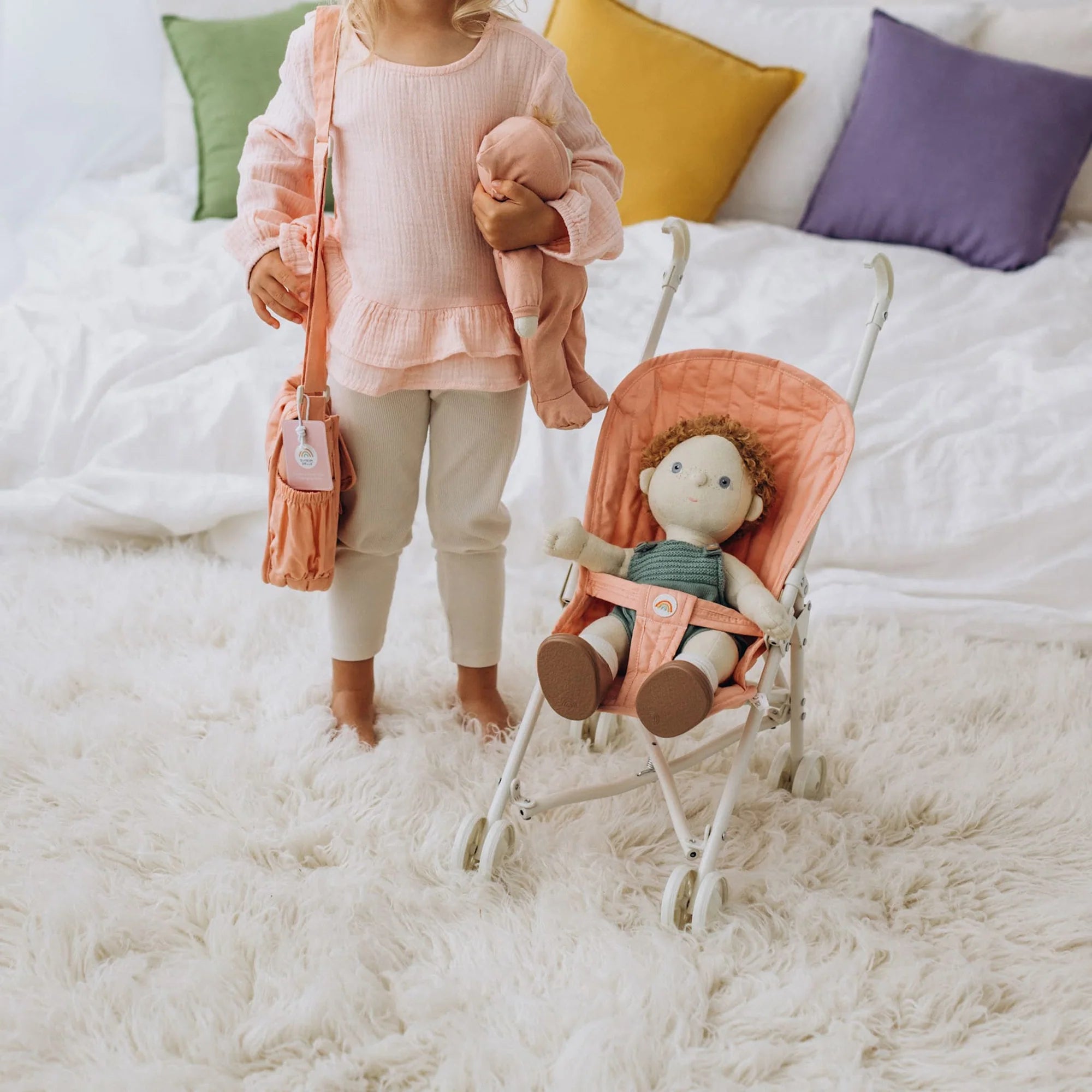 A child stands on a fluffy white rug, wearing a pink top and holding a soft doll. Beside them is the OLLI ELLA Sollie Stroller Rose with another doll comfortably seated inside. The background shows a white bed decorated with colorful pillows.