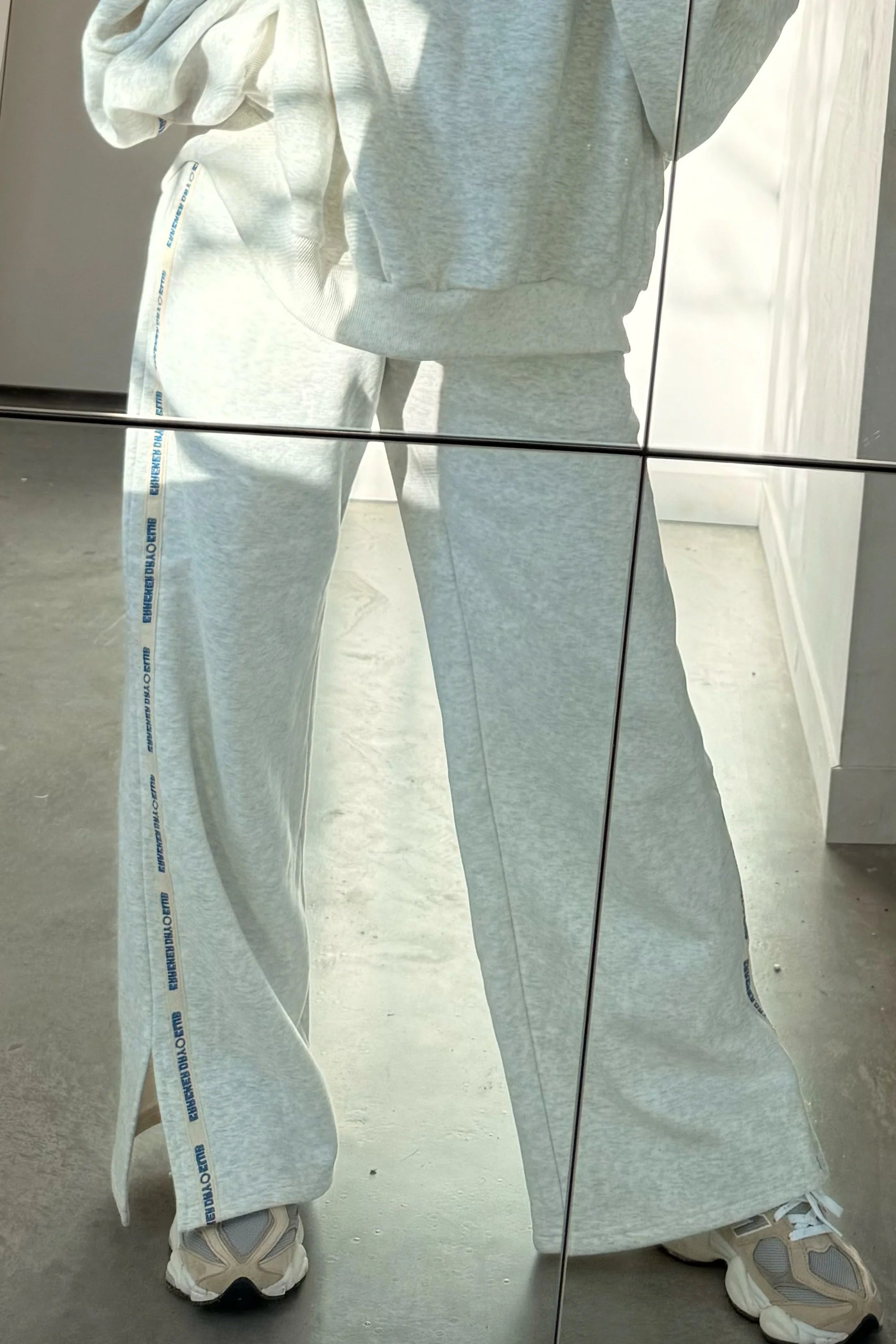 A person wears CRACKER DAY CLUB’s Cloud Racer Sweatpants in Grey Marle with text detailing and a matching sweatshirt, standing indoors on a gray floor. Striking horizontal and vertical reflections appear in the mirror photo.