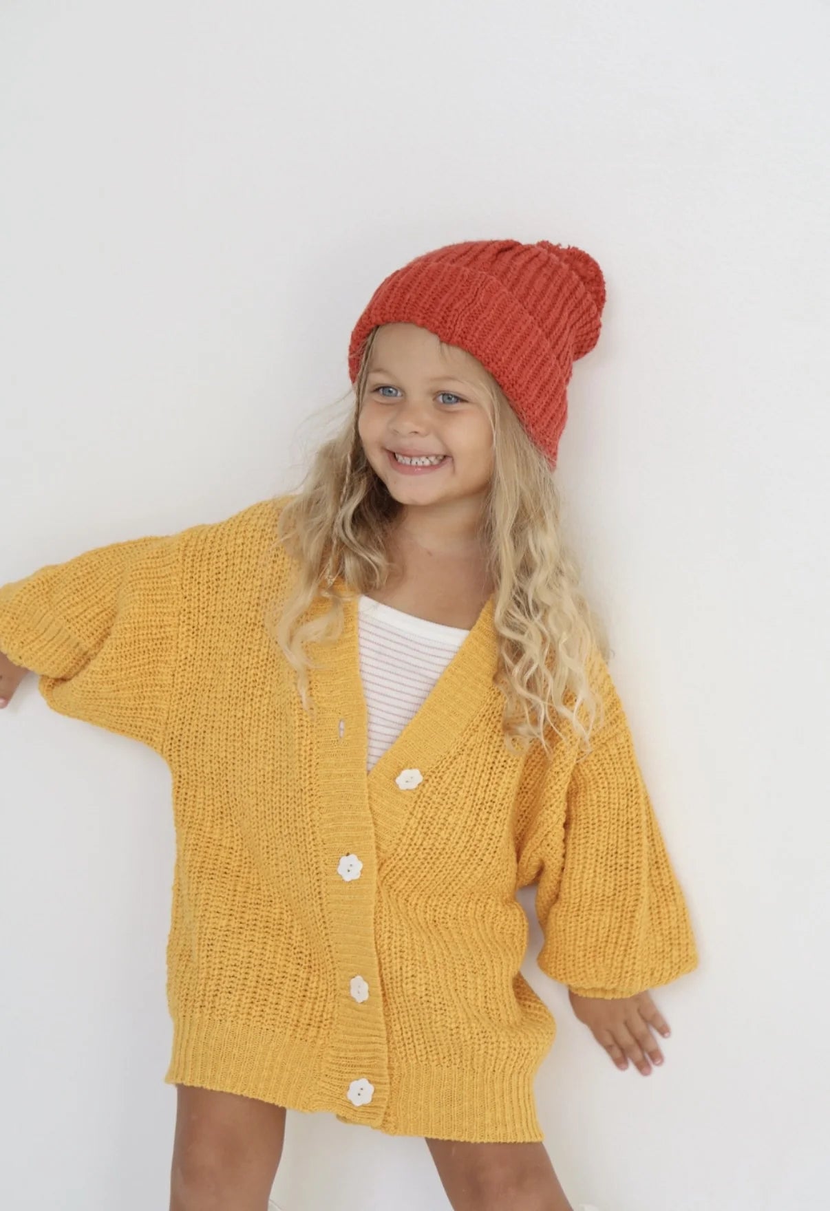 A smiling young child with long blonde hair wears the EN. THE LABEL En The Label - Knit Beanie Red Wine and a cozy white outfit, standing against a plain white background—the perfect kids' winter accessory.