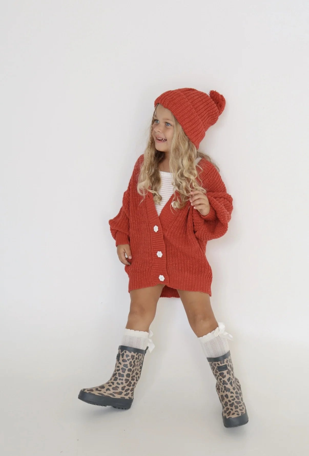 A young girl with long blond hair wears the EN. THE LABEL "En The Label - Knit Beanie Red Wine," paired with a matching oversized cardigan, white outfit, and leopard-print rain boots, smiling against a plain white background.