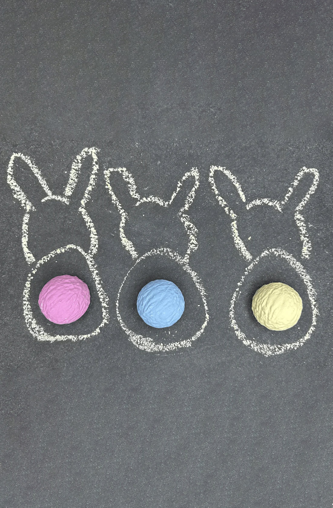 Three simple bunny shapes are drawn in white TWEE chalk on a dark gray surface, each with a colored belly. The left bunny has a pink belly, the middle bunny has a blue belly, and the right bunny has a yellow belly. This limited edition Bunnytails Sidewalk Chalk set makes for a delightful gift for little ones using non-toxic washable chalk.