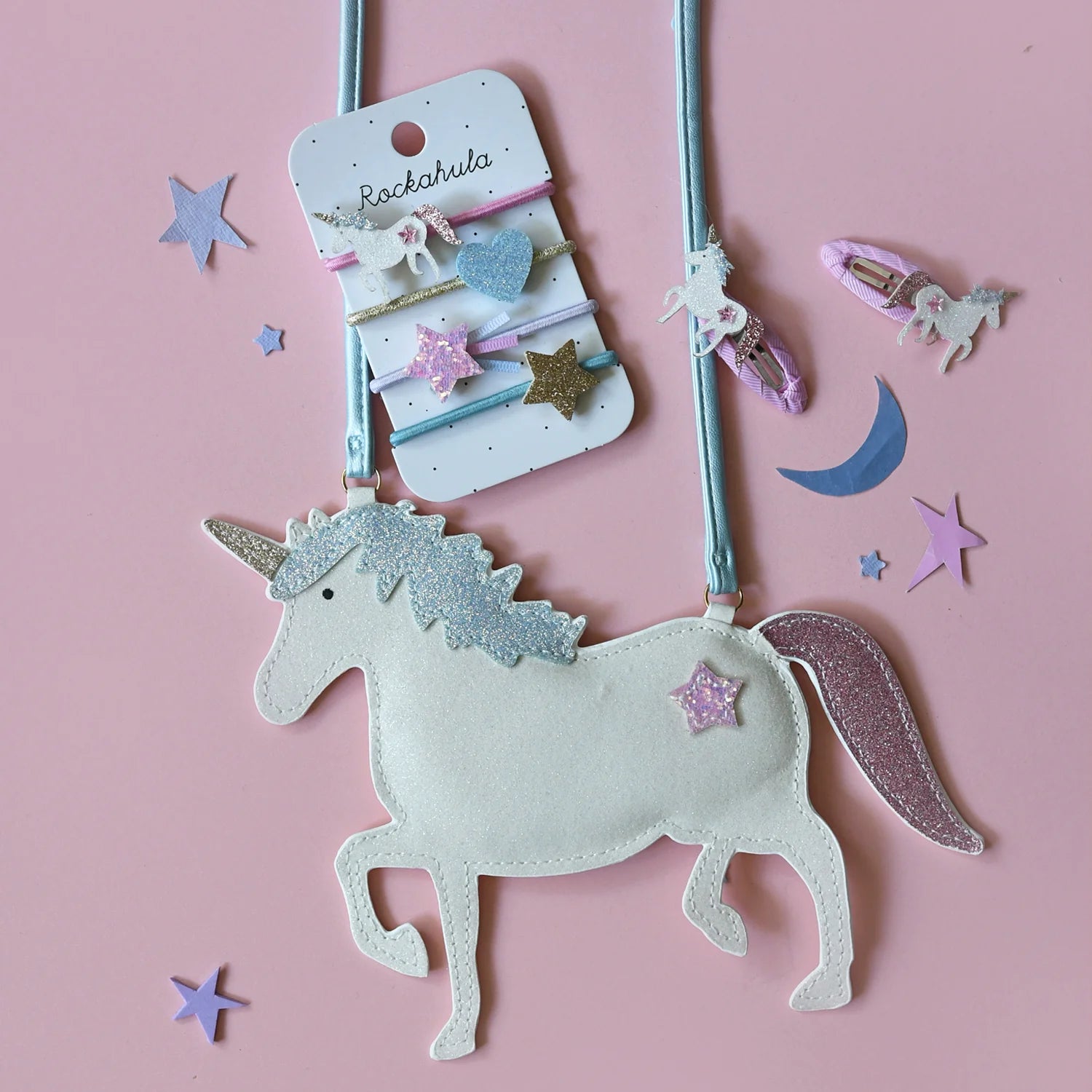 Flat lay of pink and pastel unicorn-themed accessories on a pink background, featuring the ROCKAHULA Rockahula - Unicorn Glitter Bag with a metallic blue strap, star and moon decorations, whimsical hairbands, coin pouch, and small unicorn figurines.