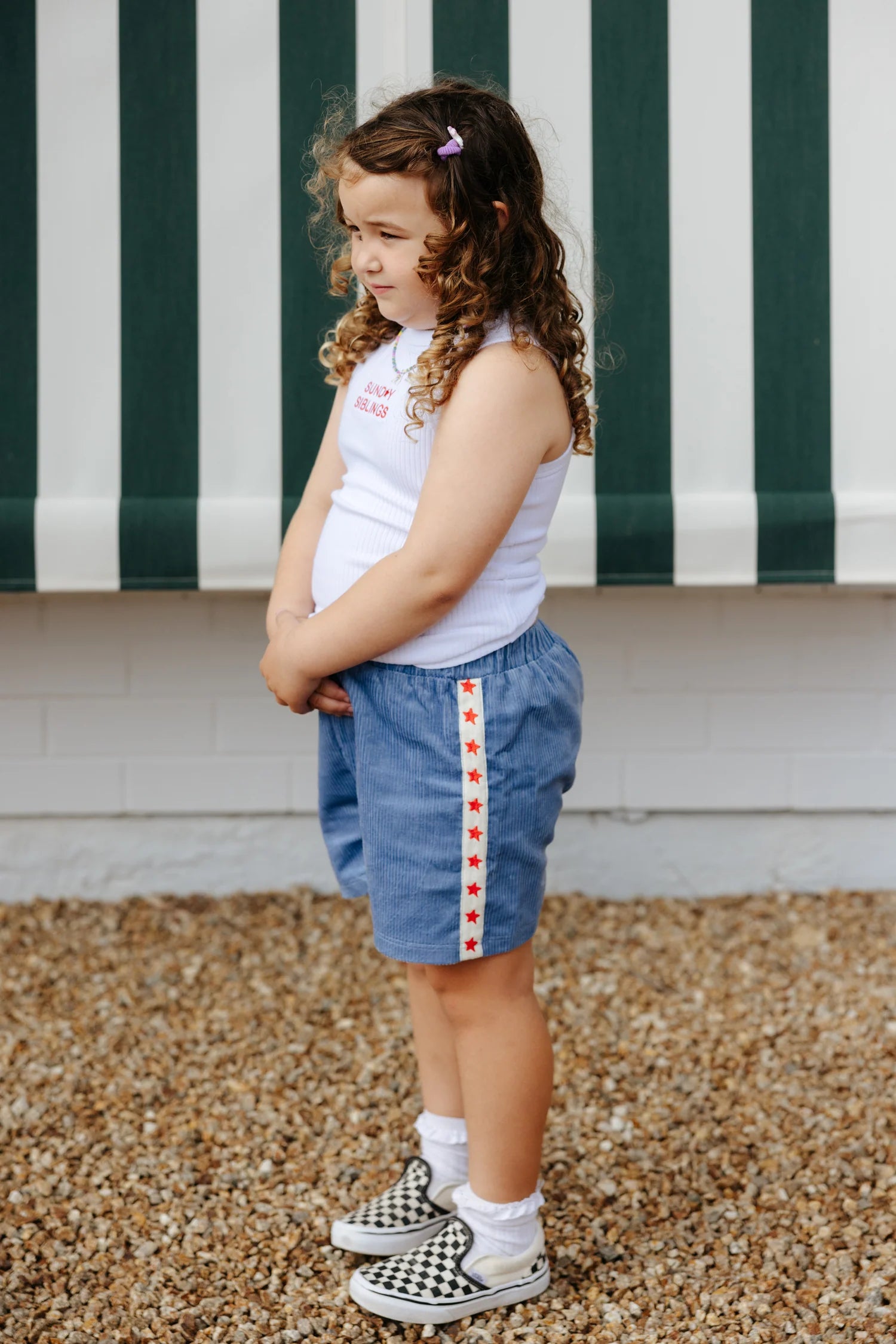 A young child stands on gravel, looking away from the camera. They sport a white tank top and the Playtime Cord Shorts Blue by SUNDAY SIBLINGS, adorned with a red star stripe. Checkered slip-on shoes complete the look, while curly hair held by a purple clip adds charm. A striped awning provides shade in the background.