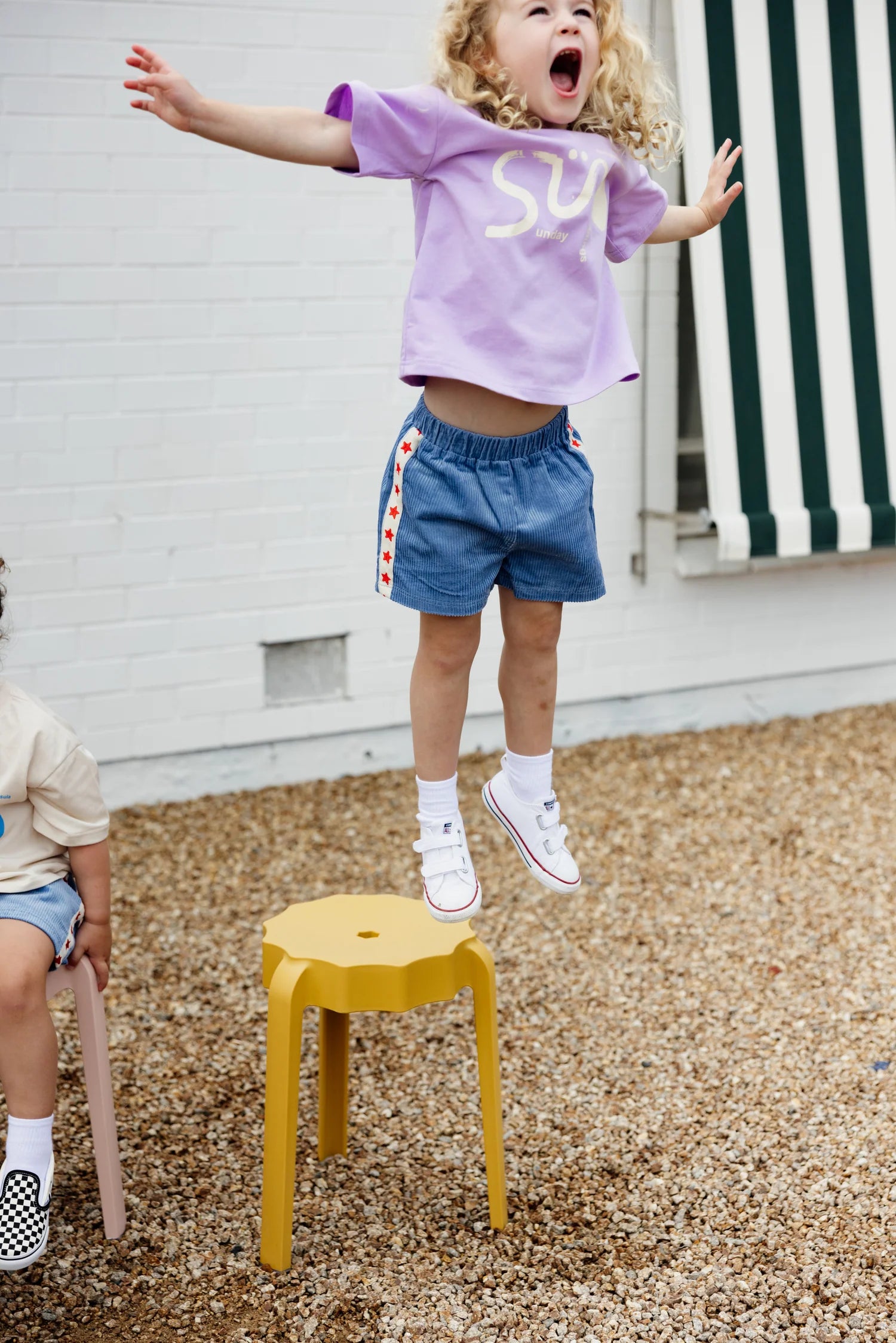 A child with curly hair, wearing a purple shirt and SUNDAY SIBLINGS Playtime Cord Shorts Blue featuring star embroidery, is joyfully jumping off a yellow stool on a gravel surface. The backdrop includes a white brick wall and striped awning. Another child is partially visible, sitting on a stool.