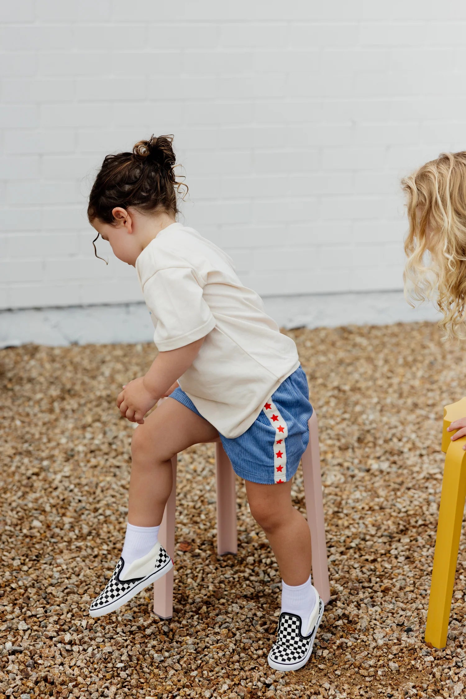 A child with short hair climbs onto a pink stool, wearing a beige t-shirt and blue Playtime Cord Shorts by SUNDAY SIBLINGS that feature a red star stripe, white socks, and checkered slip-on shoes. Nearby, another child with long curly hair sits on a yellow chair. They are on a gravel surface.