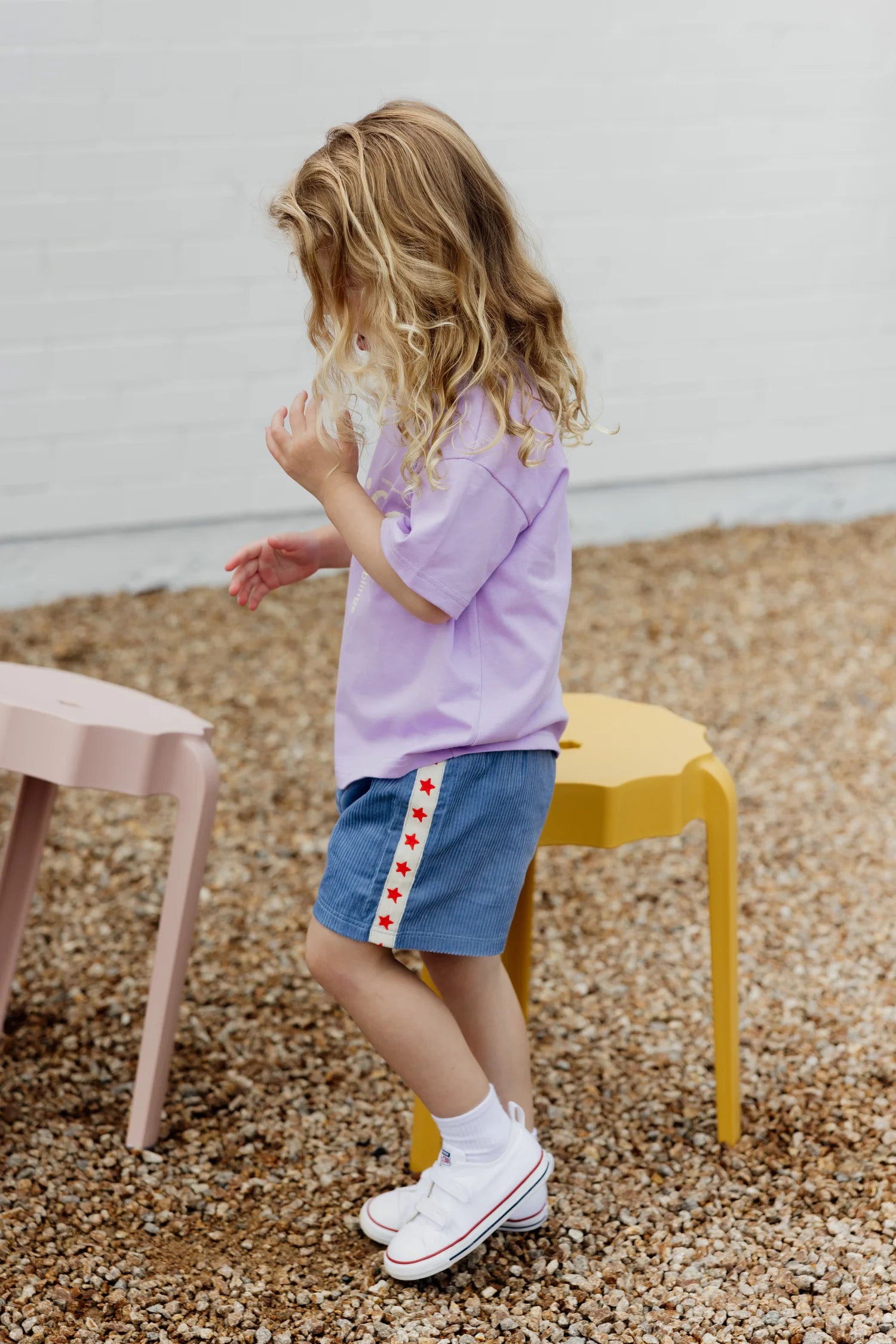 A young child with long blonde hair stands outdoors on gravel, dressed in a lavender shirt and SUNDAY SIBLINGS' Playtime Cord Shorts in blue, featuring star embroidery. White socks and sneakers complete the look as they stand beside pastel-colored outdoor stools, ready for playtime.