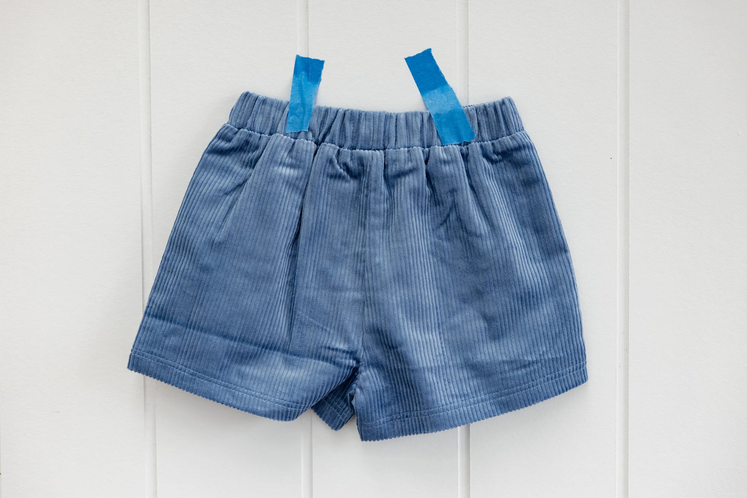 The Playtime Cord Shorts Blue by SUNDAY SIBLINGS are displayed on a white paneled wall, secured with two strips of matching tape. These shorts, made from blue cord fabric, feature an elastic waistband and front pleats. Subtle star embroidery adds a playful touch to their design.