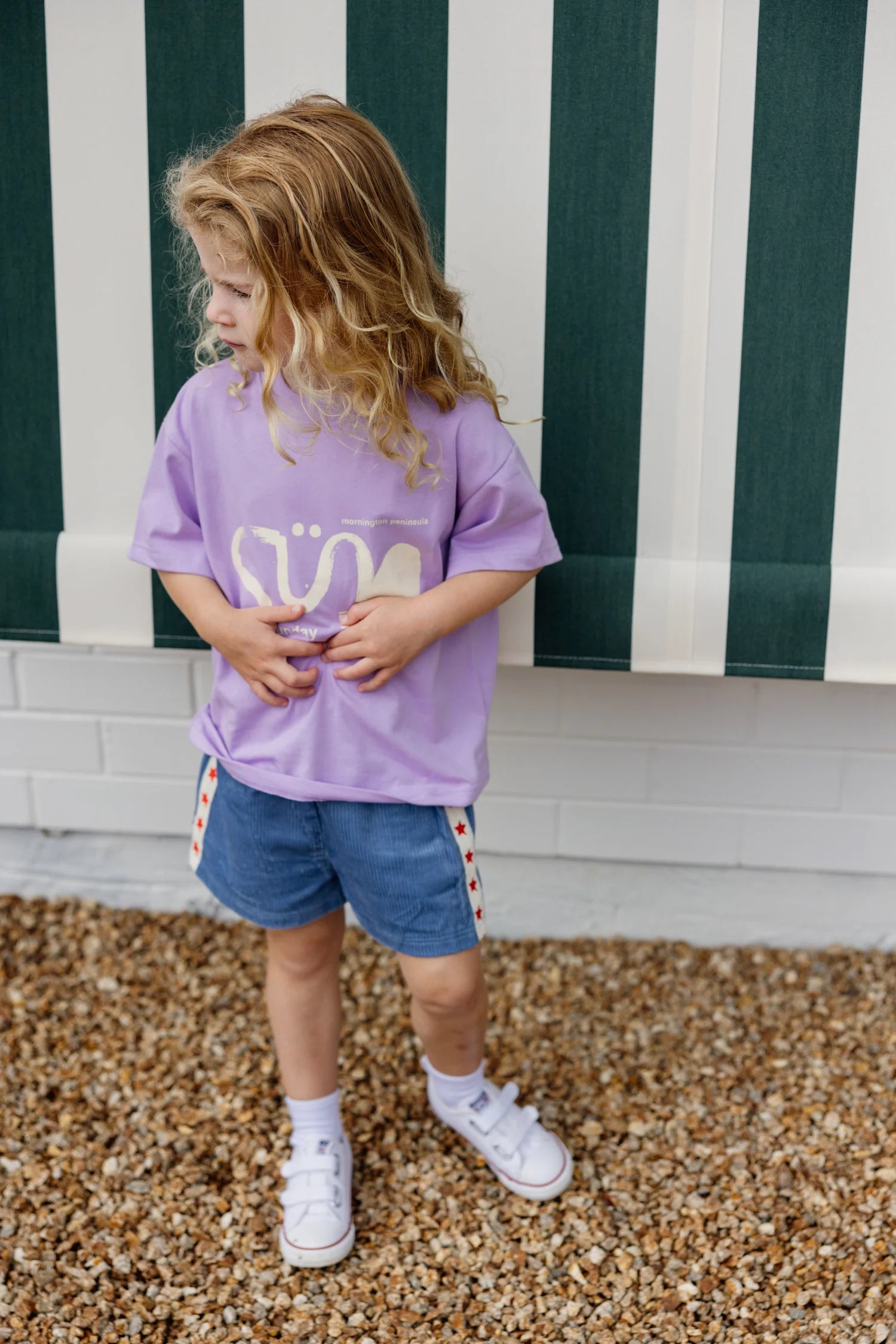 A young child with long blonde hair stands on a gravel path, wearing a purple shirt with a graphic design and SUNDAY SIBLINGS' Playtime Cord Shorts Blue. White socks peek from their sneakers, framed by the green and white striped awning against the white brick wall.