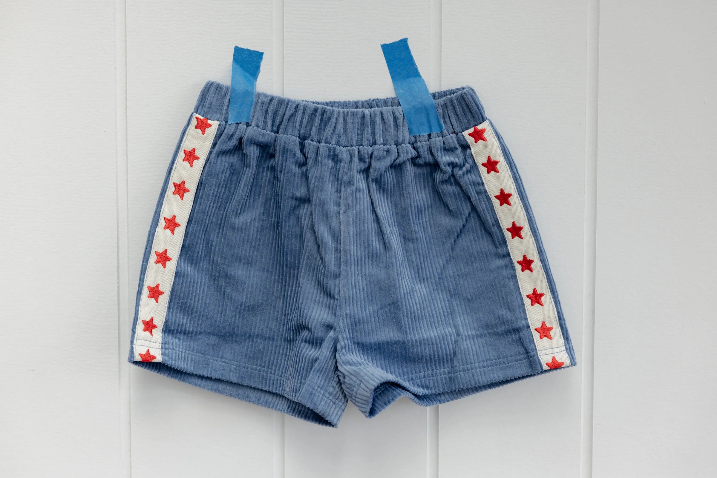 Playtime Cord Shorts Blue by SUNDAY SIBLINGS showcase white stripes with red star embroidery on each side, attached to a white wall using blue tape.