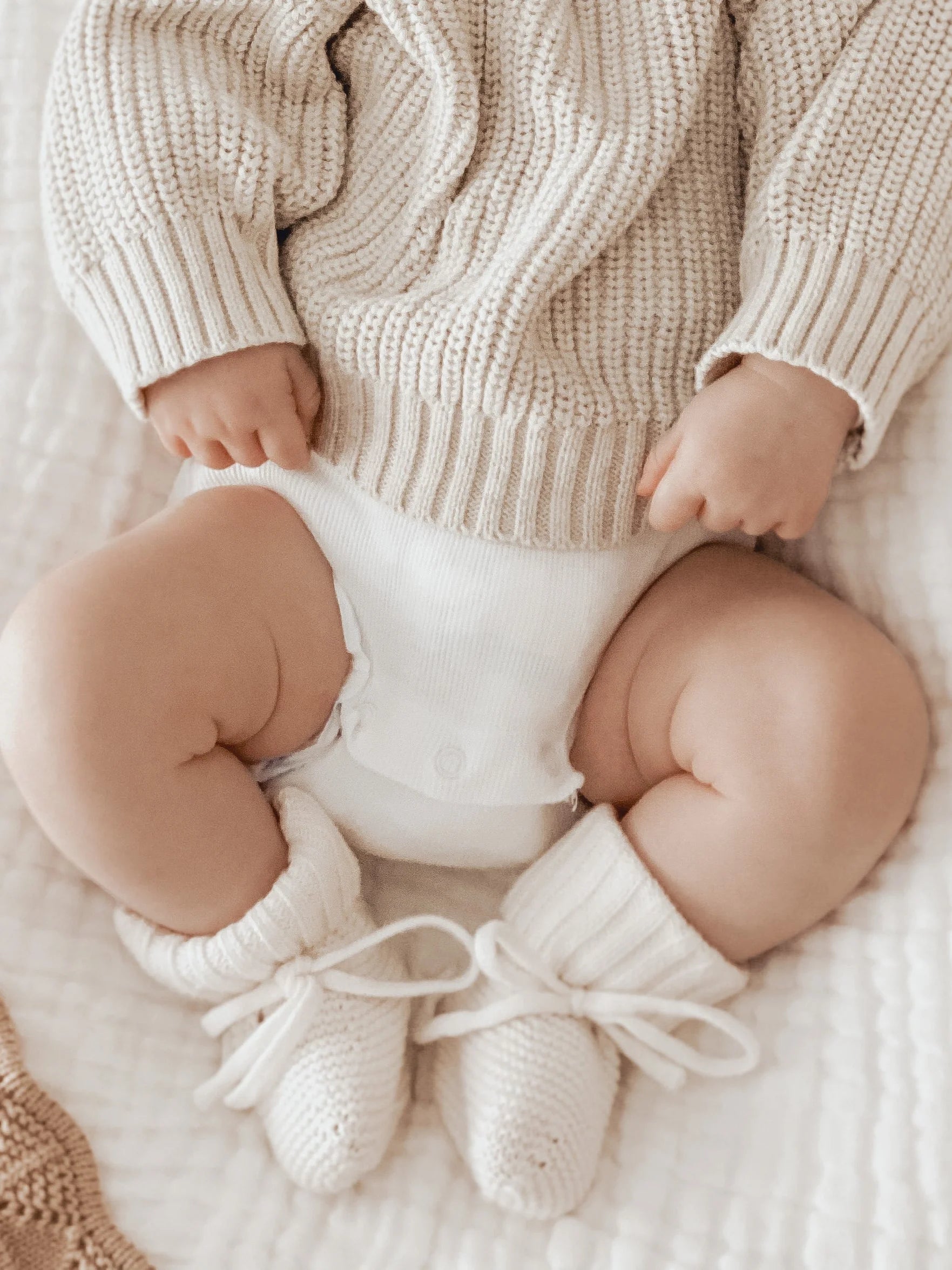 A baby in a cream knit sweater, white onesie, and ZIGGY LOU Booties Milk sits on a soft blanket, hands on the sweater and legs slightly apart.