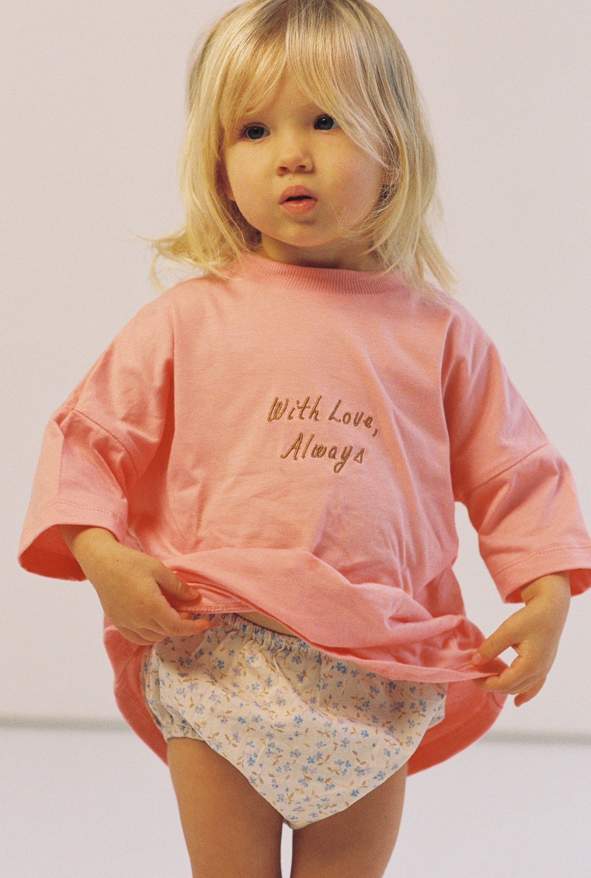 A young child with blond hair wears the Illoura the Label - With Love Tee Flamingo by ILLOURA THE LABEL and floral patterned shorts, standing against a plain background.