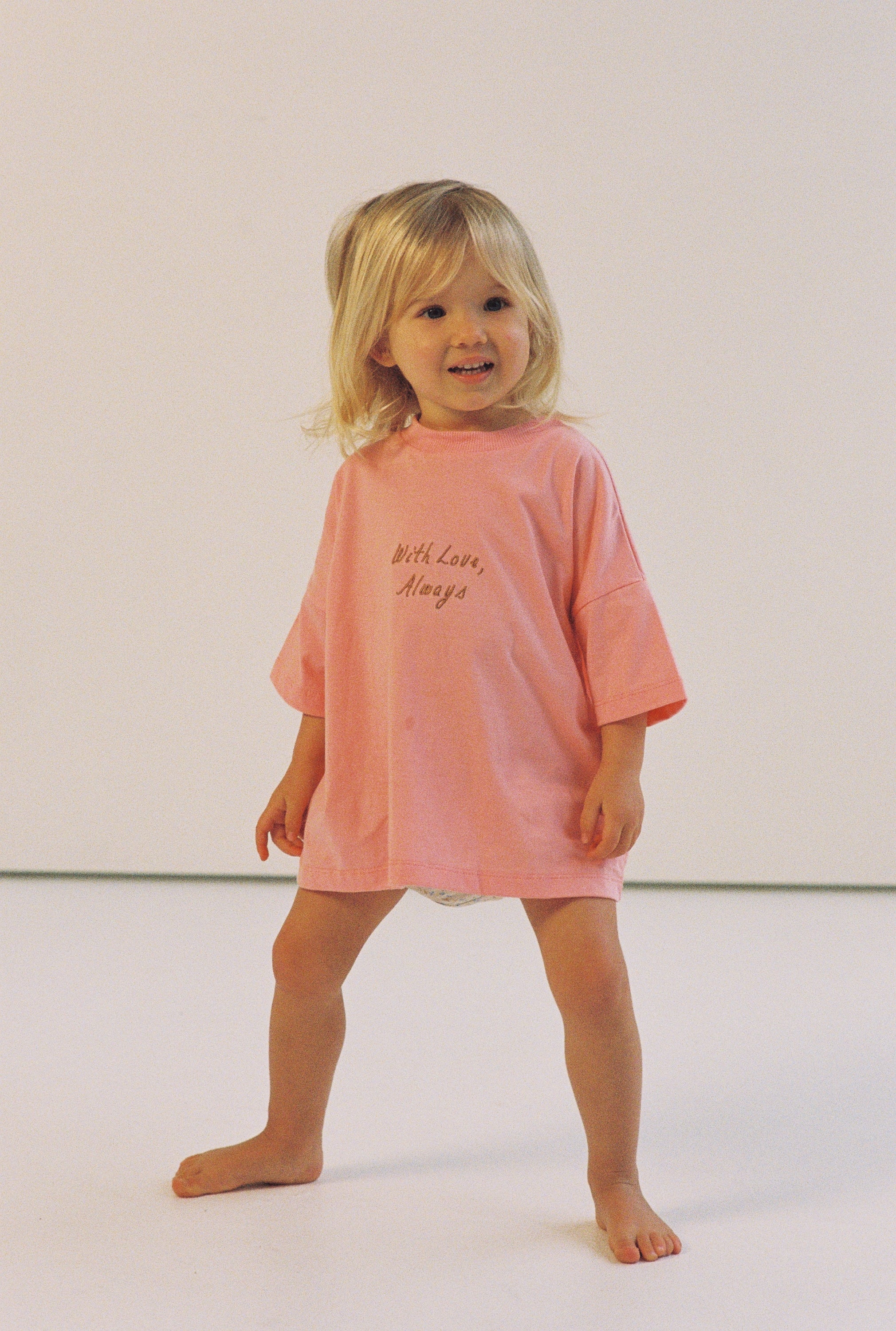A young child with blond hair stands barefoot and smiles slightly in the Illoura the Label - With Love Tee Flamingo by ILLOURA THE LABEL, set against a plain white background.