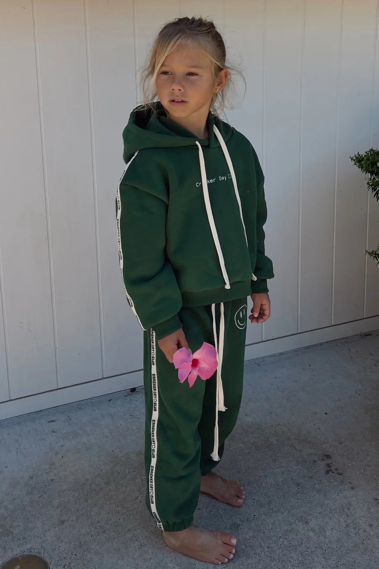 A young girl stands barefoot on concrete, wearing the CRACKER DAY CLUB Cracker Day Club - Kids Cloud Racer Hoodie Forest and matching sweatpants with white stripes, holding a pink flower by a white wall and green bush.