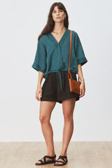 Woman wearing Eda Blouse in Seagrass with black shorts and tan bag