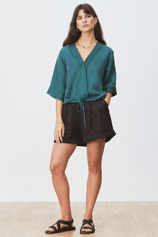 Front view of woman wearing Eda Blouse in Seagrass with black shorts