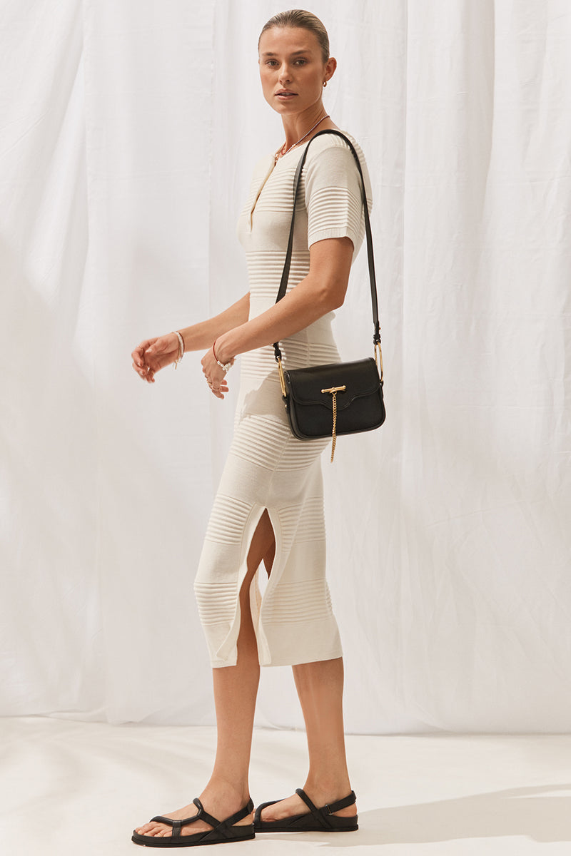 Woman wearing Isabella Dress in Off White with black handbag. Full length, side view.